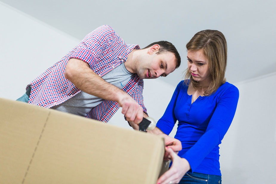 Muškarac i žena zajedno zatvaraju kartonsku kutiju, muškarac koristi selotejp, dok žena pažljivo gleda. Oboje su fokusirani na pakovanje i izgledaju kao da se pripremaju za hitnu selidbu.